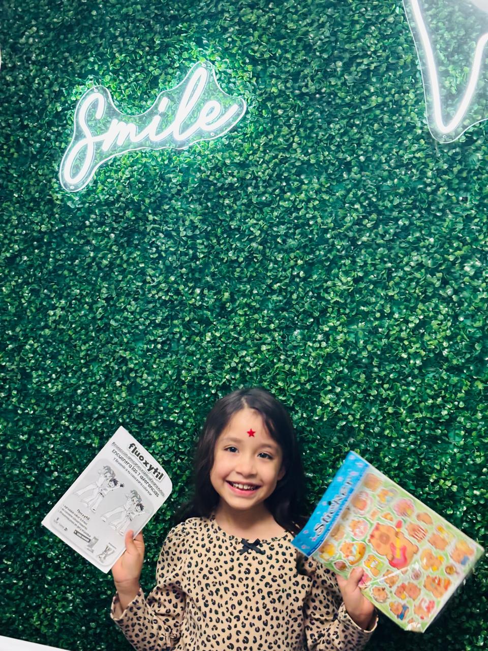Niña con premio y stickers frente al muro verde con letrero Smile neón en consultorio dental