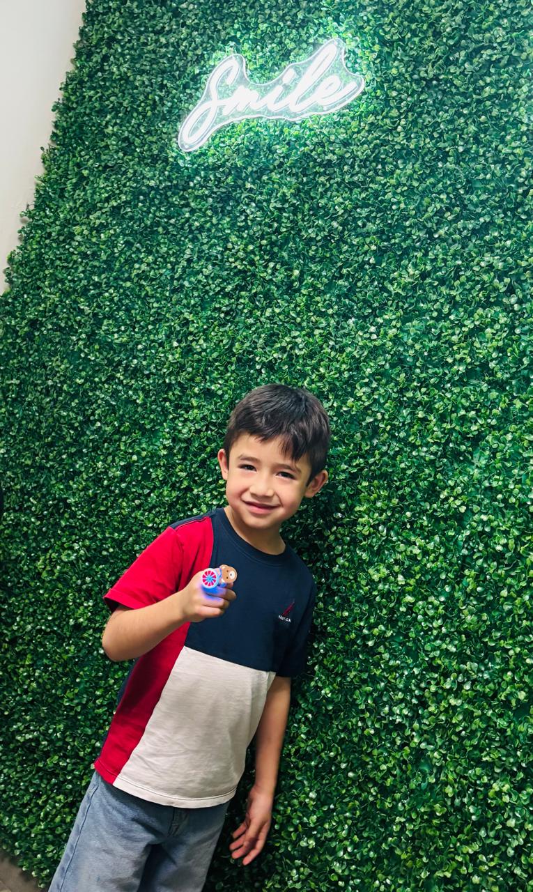 Niño con sticker de premio frente al muro verde con letrero Smile neón en Especialidades Dentales